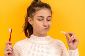 A girl is holding a toothbrush and clear aligners at her eye level and looking at them